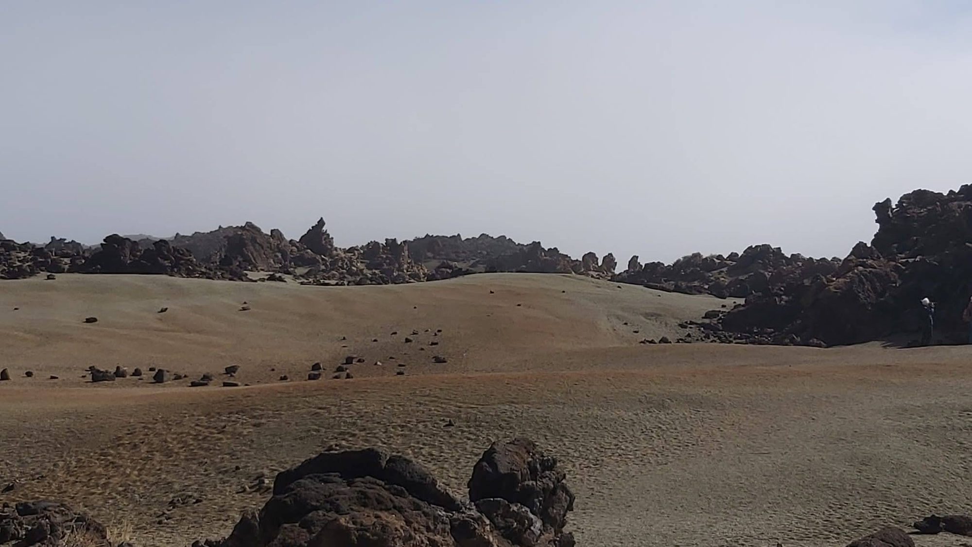 Paisaje volcánico árido con formaciones rocosas oscuras y terreno arenoso bajo cielo claro.